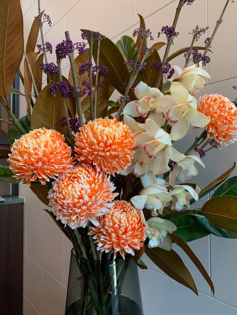 A tall, elegant floral arrangement featuring vibrant orange pom-pom chrysanthemums with densely petaled blooms, complemented by creamy white orchids with pink accents on their petals and elongated ste