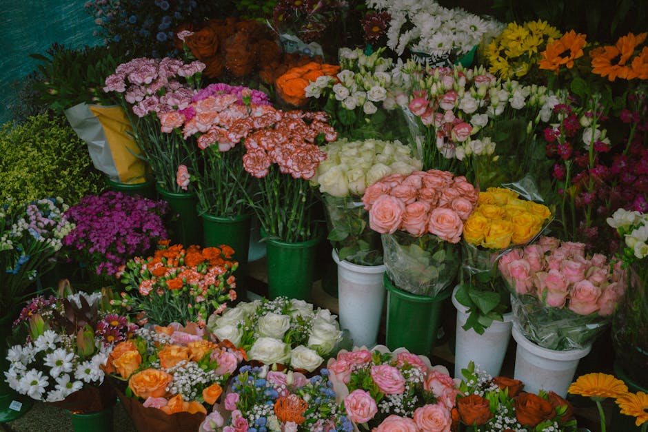 A vibrant display of fresh flower arrangements and bouquets arranged in green and white pots and wrapping, featuring a variety of blossoms including pink roses, white roses, yellow roses, orange bloss