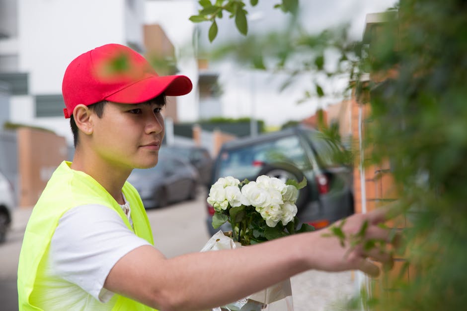 A young male florist wearing a red cap and a high-visibility yellow vest is carefully arranging a bouquet of white hydrangeas outdoors, with a background of parked cars, residential buildings, and gre