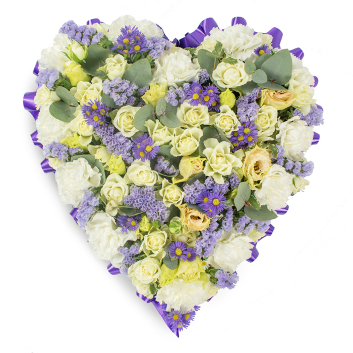 A lovingly composed heart of flowers set against a plain white studio backdrop, where every element is chosen to read clearly from a distance and comfort up close. The heart's edge is defined by a rich, looped purple satin ribbon that runs continuously around the perimeter, its neat folds creating a composed frame for the blooms within. Within that border, pale creams and buttery yellows dominate: clusters of cream and pale yellow roses-some in tight bud form, others with petals easing outward-sit densely packed to produce a soft, layered texture, while generous tufts of white carnations add classical volume and a reassuring, familiar presence. Interspersed are clouds of lavender statice and lively purple, daisy-like asters with vivid yellow centres, which introduce points of deeper colour and a gentle, rhythmic contrast across the surface. Blue-green eucalyptus leaves are threaded through the design, their rounded silhouettes and cool tones giving shape and a subtle botanical punctuation that calms the palette. The lighting is even and soft, highlighting the velvet of rose petals, the frill of carnations, and the papery feel of statice; the overall impression is one of delicate craft rather than extravagance. This arrangement, arranged with both precision and tenderness, suits funeral services, wakes, or graveside ceremonies-an outward symbol of steady affection and remembrance. Locally resonant for families in Abbey Wood and nearby Plumstead or Belvedere, the piece conveys sympathy and respect in quiet, reassuring language, offering a dignified floral embrace for those saying goodbye.